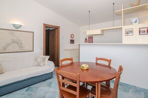 Een eetgedeelte met een ronde houten tafel en stoelen, een bank, een wandplank met ingelijste foto's en een deur naar een andere kamer.