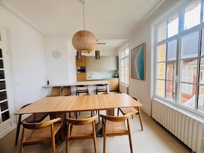 A dining area with a wooden table and chairs, a hanging light fixture, and a view through large windows.