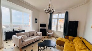 Ein Wohnzimmer mit einem beigen Sofa, einem gelben Sofa, einem schwarzen Tisch, einem weißen Sessel und einem Fernseher an der Wand.