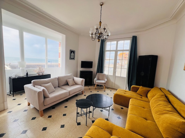 A living room with a beige sofa, a yellow sofa, a black table, a white armchair, and a TV on the wall.