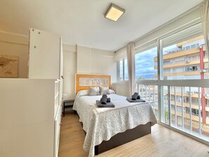 A bedroom with a bed, a headboard with a relief, a window with a view of buildings, and a ceiling light. - Terrasol EL Cisne Torre DEL MAR (Velez-Malaga)