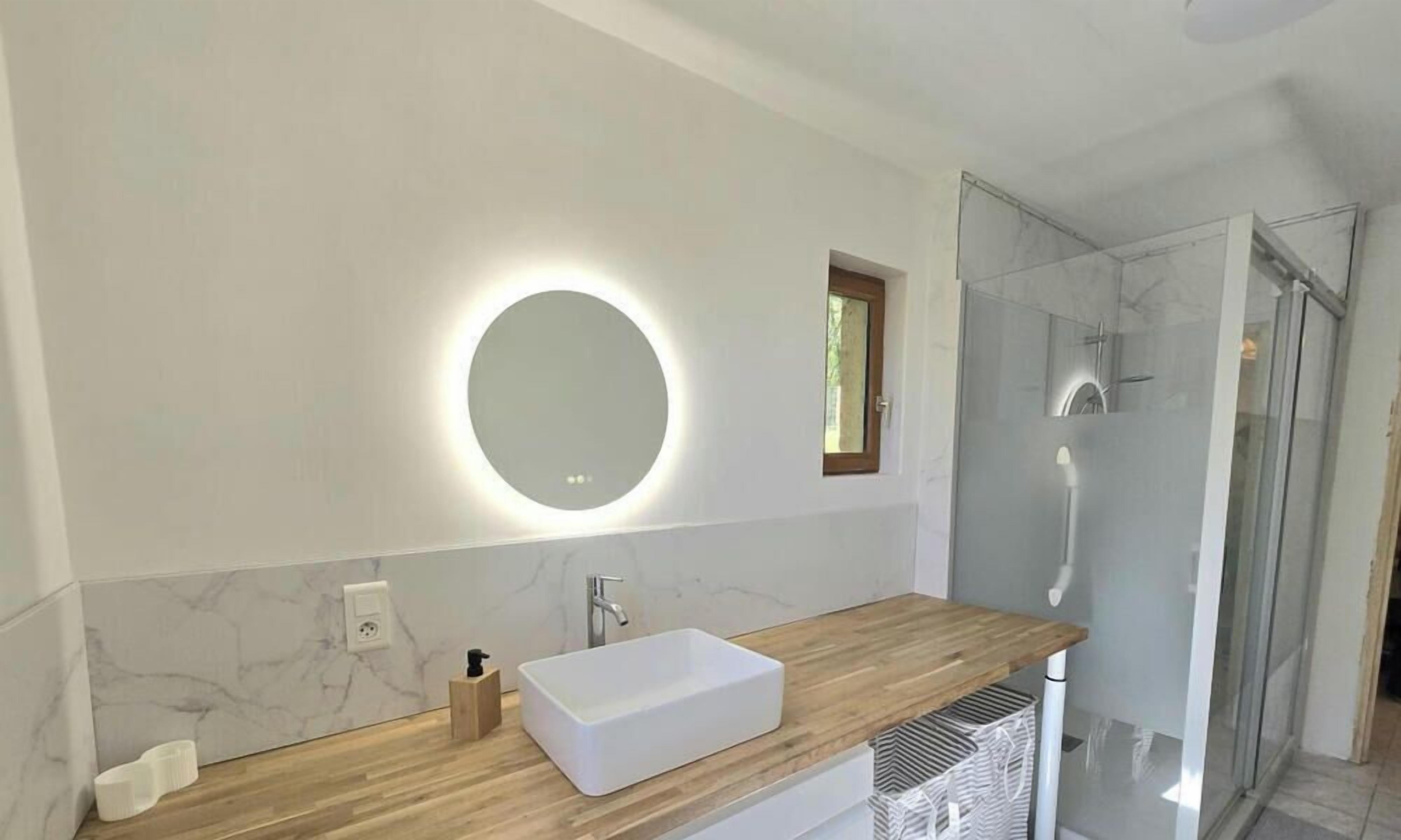 Une salle de bain moderne avec un meuble-lavabo en bois, un lavabo blanc, un miroir rond avec éclairage intégré, une douche avec des portes en verre et des murs en marbre.