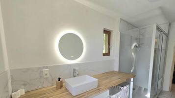 Une salle de bain moderne avec un meuble-lavabo en bois, un lavabo blanc, un miroir rond avec éclairage intégré, une douche avec des portes en verre et des murs en marbre.