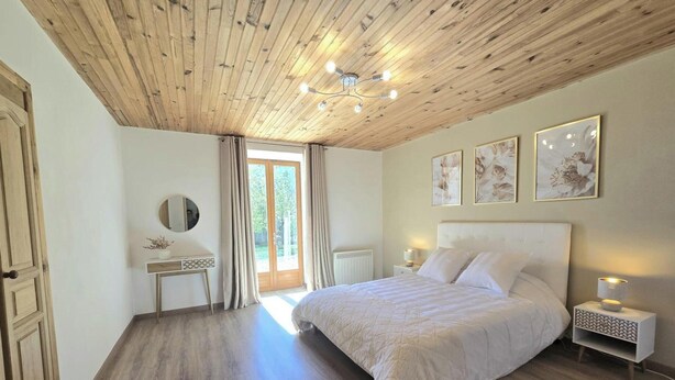Ein Schlafzimmer mit Holzbalkendecke, einem Bett mit weißer Bettwäsche, einem Nachttisch mit Lampe und einem Fenster mit Vorhängen.