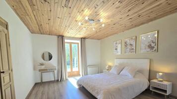 Une chambre avec un plafond en bois, un lit recouvert de draps blancs, une table de chevet avec une lampe et une fenêtre avec des rideaux.