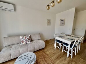 A modern living room with a sofa, a dining table with chairs, a small round table, and a ceiling light fixture. - Air-conditioned apartment, balcony - 4 guests (Le Lavandou)