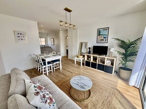 A modern living room with a sofa, a dining table, a TV, and a kitchen in the background. - Air-conditioned apartment, balcony - 4 guests (Le Lavandou)