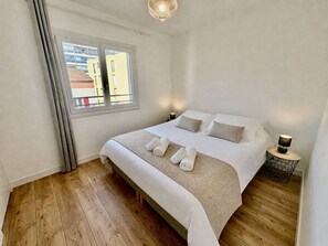 A hotel room with a large bed, a window with curtains, a bedside table with a lamp, and a view of buildings outside. - Air-conditioned apartment, balcony - 4 guests (Le Lavandou)