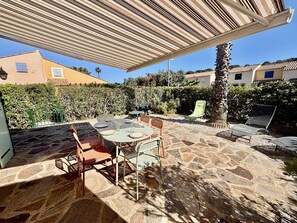 Eine Terrasse im Freien mit Steinboden, einem Tisch und Stühlen, einer Pergola darüber und Blick auf eine Wohngegend mit Häusern und Grünflächen.