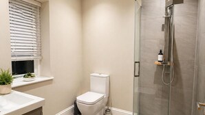 A modern bathroom with a toilet, a glass shower enclosure, and a window with blinds. - Modern Cottage Sleeps 4 With Patio (Royston)