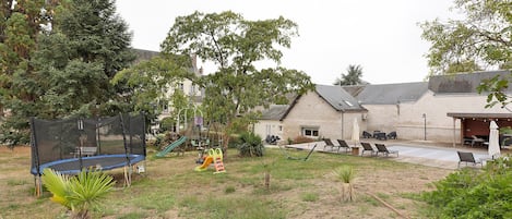 Un jardin à l’arrière d’une maison, aménagé avec un trampoline, un ensemble de jeux pour enfants et un espace salon extérieur.