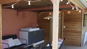 A covered outdoor area with a wooden deck, a sink, a cabinet, a sofa, and two chairs.