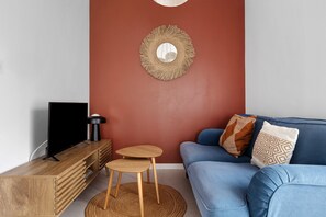 A living room with a blue sofa, a television on a wooden stand, a woven wall hanging, and a small wooden stool. - Gite "La Glycine" for 2 with Heated Pool from Mid-May to September (Veuzain-sur-Loire)
