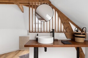 A bathroom with a wooden vanity, a round mirror, a black faucet, a white sink, a woven basket, and a leopard print pillow. - Gite "La Glycine" for 2 with Heated Pool from Mid-May to September (Veuzain-sur-Loire)
