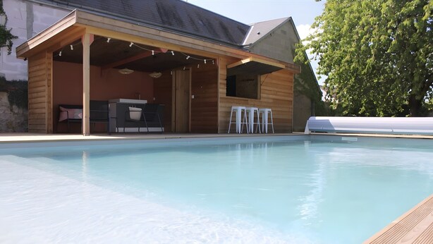 A pool with a wooden deck, a covered outdoor seating area, and a building with a sloped roof.