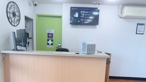 Un mostrador de recepción con un monitor de computadora, un letrero de primeros auxilios, una pantalla digital mostrando música y un dispositivo de check-out rápido.