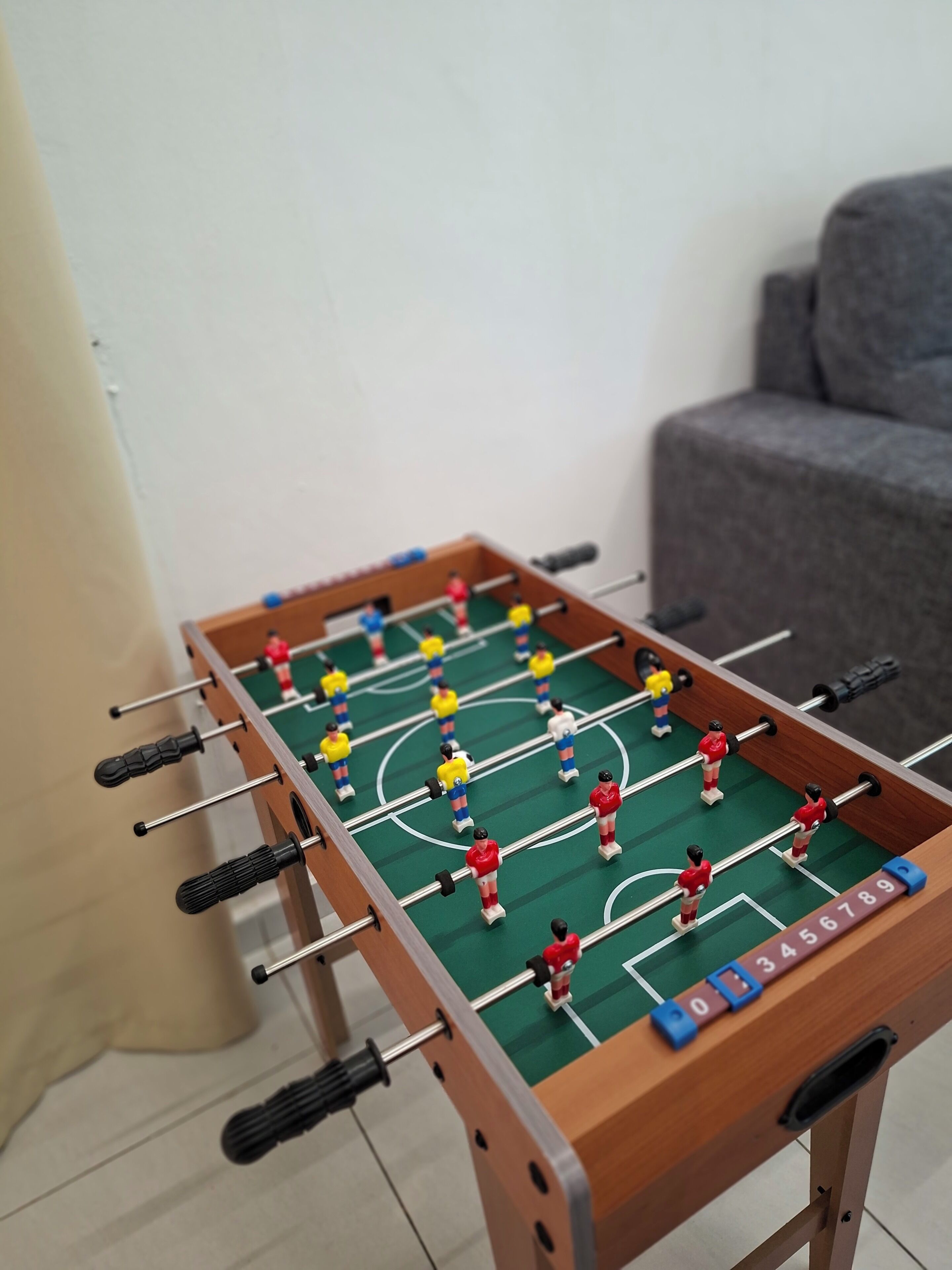 A foosball table with players in red, yellow, and blue, set against a white wall with a gray sofa in the background.