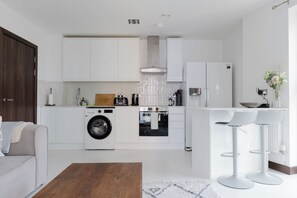 A modern kitchen with white cabinetry, a built-in washing machine, and a dining area with a white table and chairs. - Sophisticated 2BD Flat with Riverside Views (Greater London)