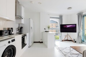 Eine moderne Küche mit Backofen, einem Bosch-Toaster und einem Kühlschrank. Eine kleine Kücheninsel beherbergt ein Spülbecken, und ein Flachbildfernseher steht auf einem Stativ. Die Wände sind weiß und ein großes Fenster ist mit Vorhängen versehen.