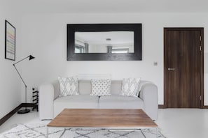 A living room with a sofa, a coffee table, a framed picture, a floor lamp, and a door. - Sophisticated 2BD Flat with Riverside Views (Greater London)