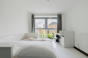 A hotel room with a bed, a desk, and a view of buildings through a window. - Sophisticated 2BD Flat with Riverside Views (Greater London)