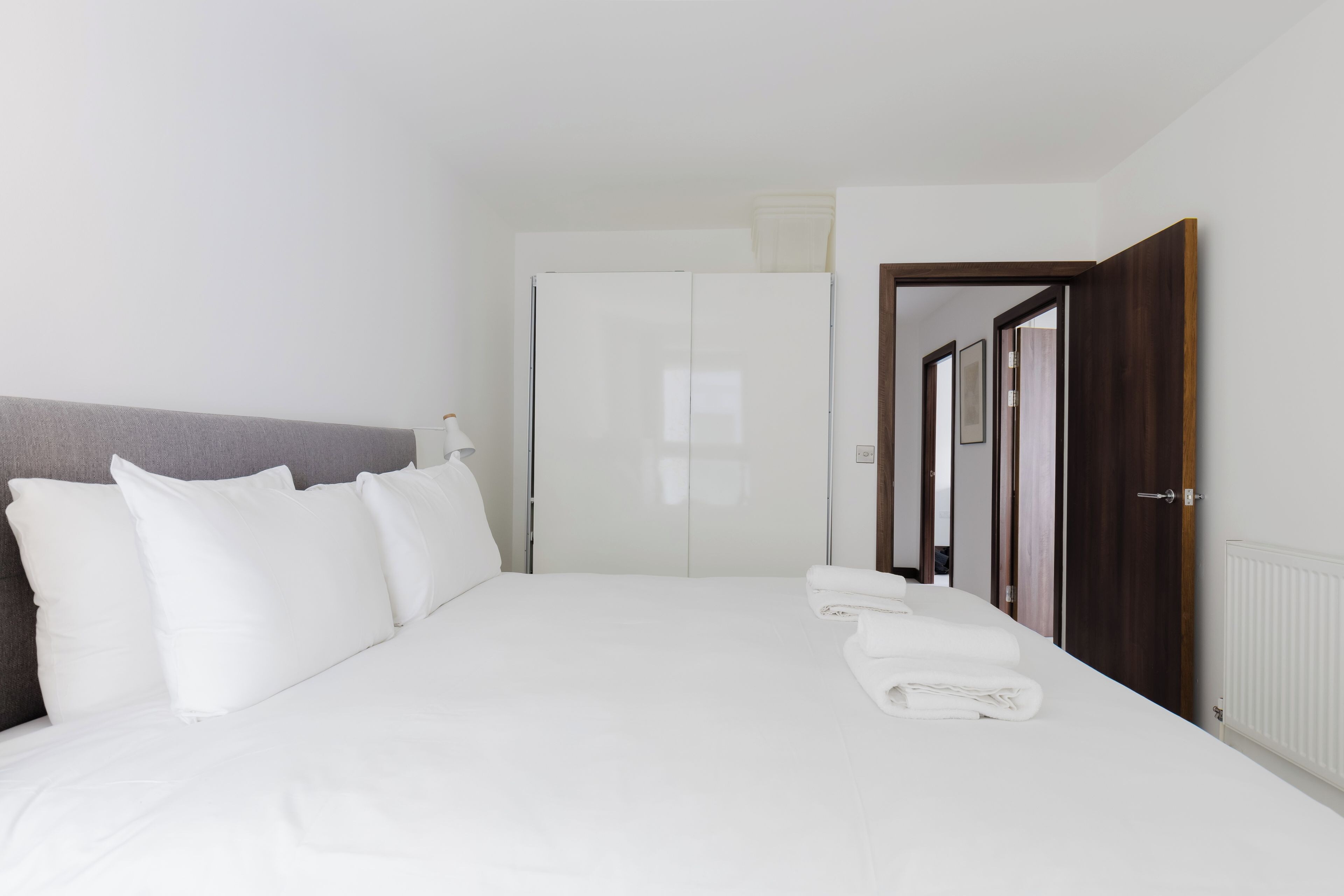 A hotel room with a bed, white linens, a headboard, a wardrobe, and a door leading to another room.