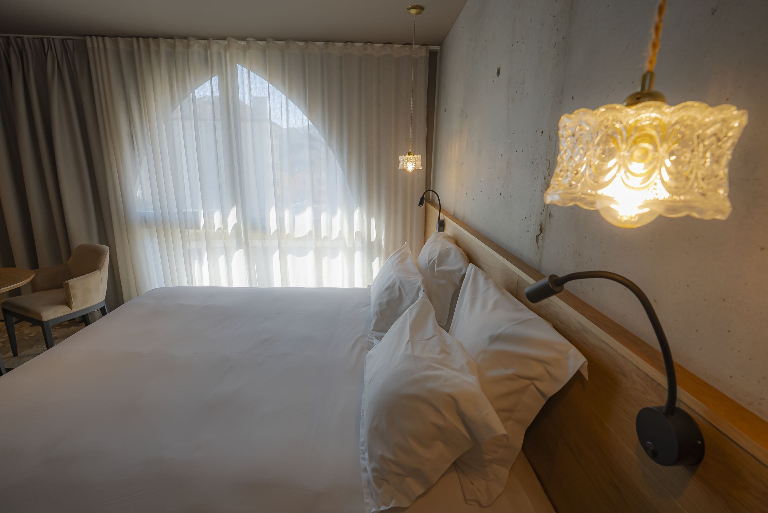 Et hotelværelse med seng, stol, lampe og et vindue med gardiner.