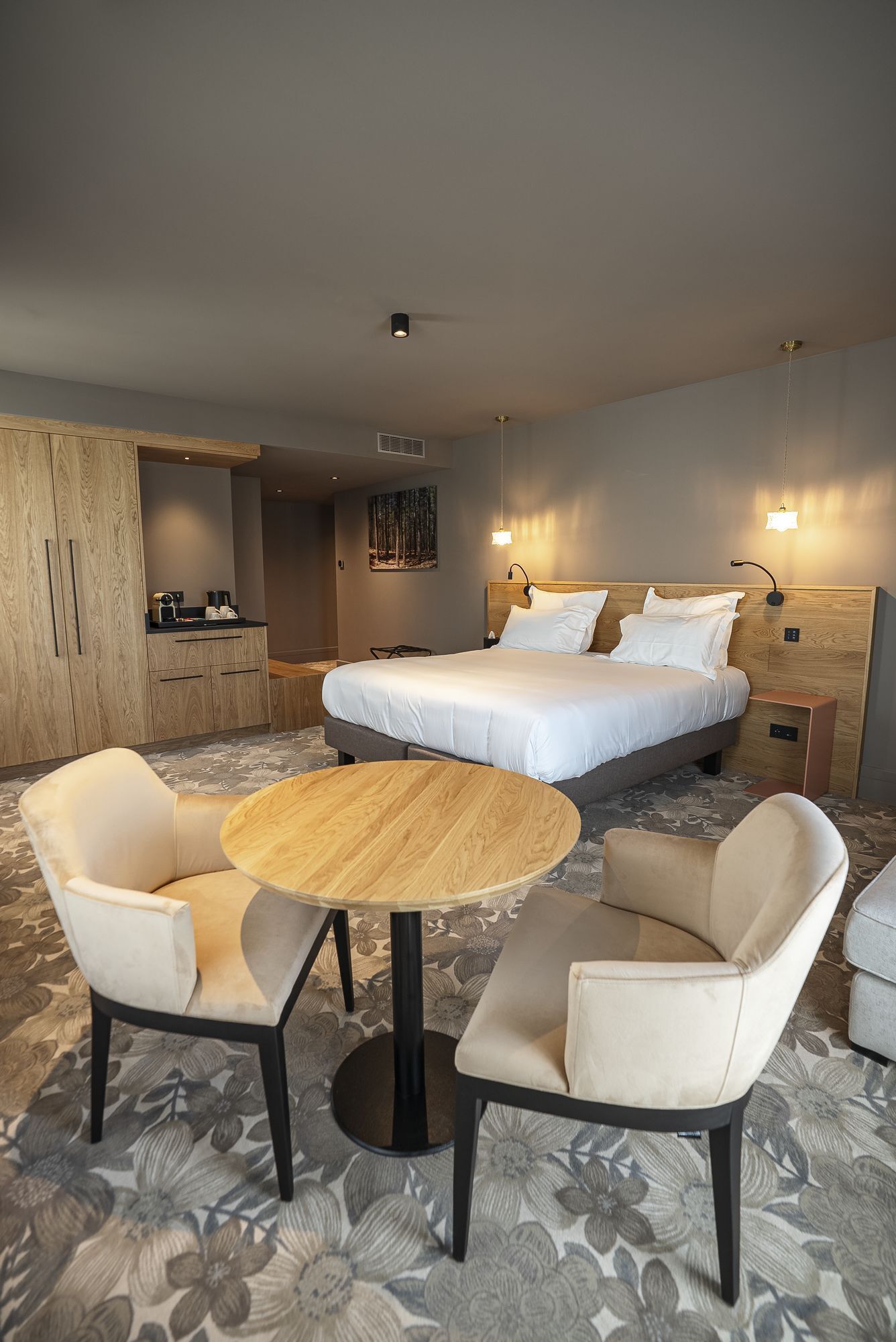 A hotel room with a bed, two armchairs, a round table, a kitchenette, and a painting on the wall.