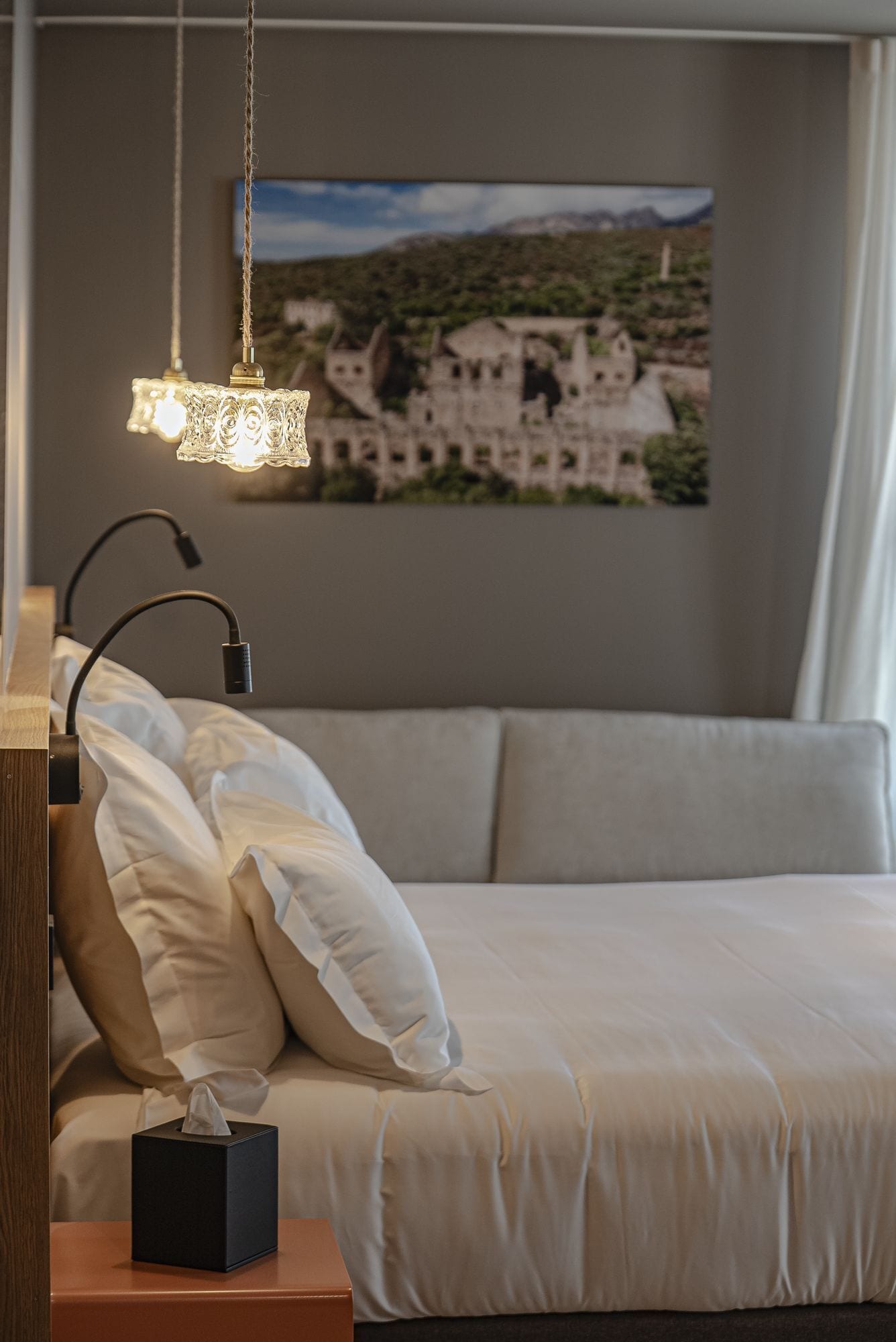Un dormitorio con cama, almohadas, una mesita de noche con una caja de pañuelos, una lámpara y un cuadro de un pueblo en la pared.