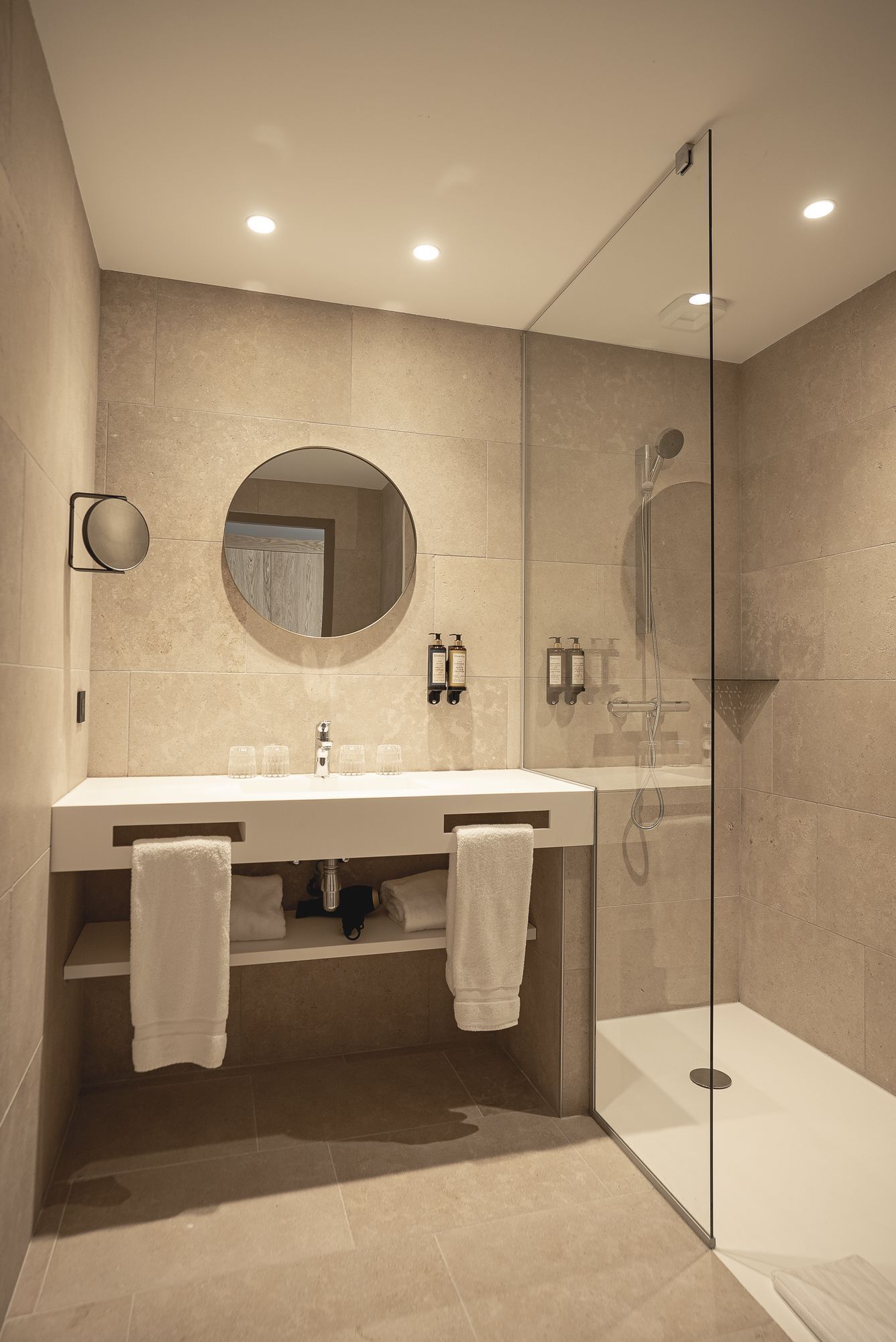 Une salle de bain moderne avec une douche à parois de verre, un miroir rond et deux lavabos avec des serviettes.