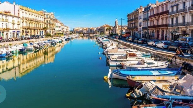 Un port de plaisance avec des bateaux amarrés le long de la rive, des bâtiments de chaque côté et un ciel dégagé.