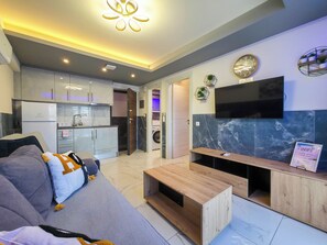 A modern living room with a sofa, coffee table, TV, and kitchen in the background. - 7 - ILEKTRA Suite by SDR (Paralia)