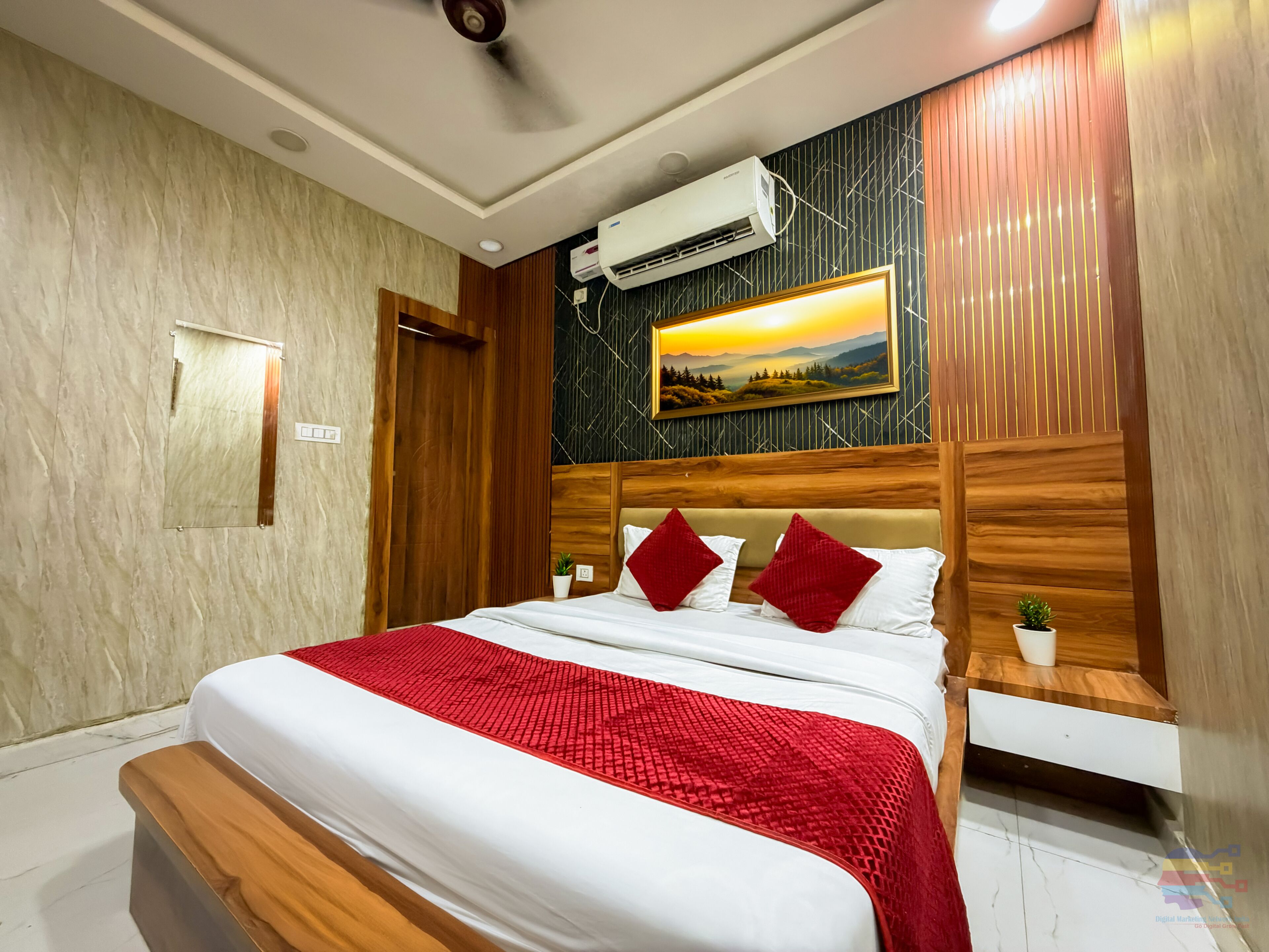 A hotel room with a bed, a wooden headboard, a wall-mounted air conditioner, a painting, and a small potted plant.