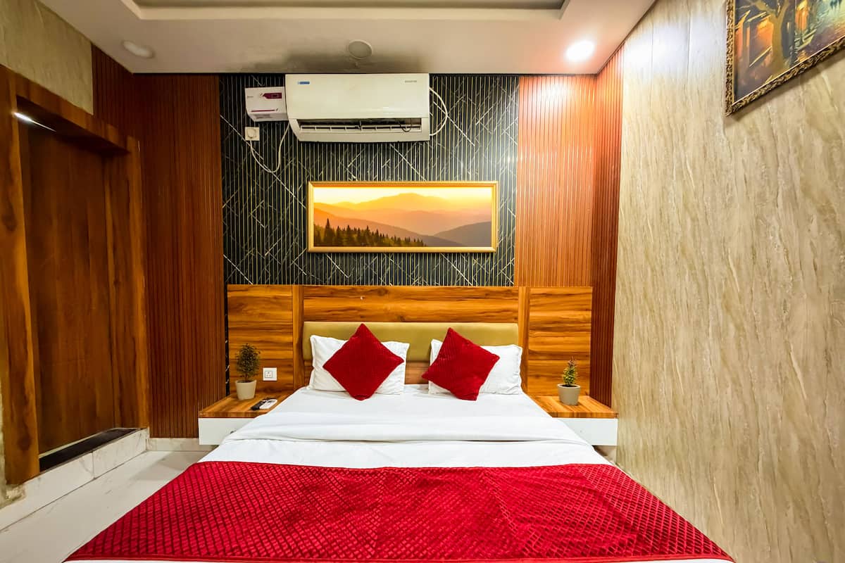 Quarto de hotel com cabeceira de madeira, cama com roupa de cama branca e detalhes vermelhos, um quadro de um pôr do sol e um ar condicionado.