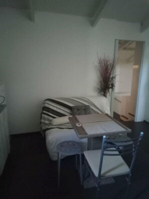 Ein kleiner, schummrig beleuchteter Raum mit einem Bett, einem Sessel, einem Tisch und einem Spiegel.