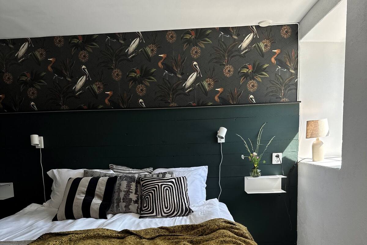 Une chambre à coucher avec un lit, une tête de lit verte, une fenêtre avec une lampe et un mur orné d’un motif représentant des oiseaux.