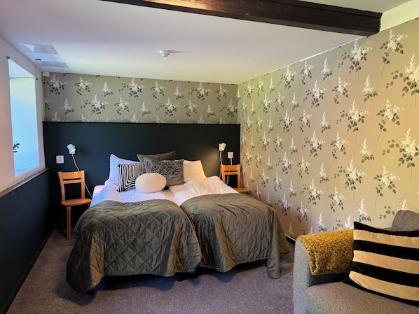 Un dormitorio con una cama, dos mesitas de noche, una mesita auxiliar, una ventana y papel tapiz floral.
