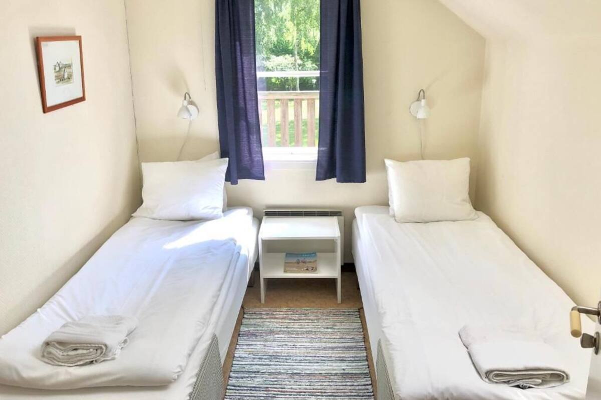 Une chambre avec deux lits simples, une petite table, une fenêtre avec des rideaux, une lampe fixée au mur et un tableau encadré.