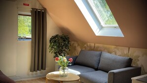 A room with a sloped ceiling, a sofa, a small table with a vase of flowers, a potted plant, and a view through a window. - Gåsen Out B&B (Burgsvik)