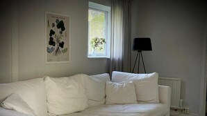 A white sofa in a room with a window, a floor lamp, and a poster on the wall. - Gåsen Out B&B (Burgsvik)