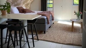A loft-style bedroom with a bed, a desk, and a chair, a dining area with a table and chairs, and a view of the outdoors through a window. - Gåsen Out B&B (Burgsvik)
