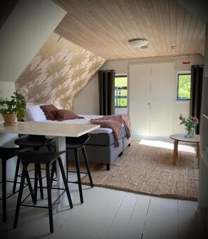 A loft-style bedroom with a bed, a desk, and a chair, a dining area with a table and chairs, and a view of the outdoors through a window.