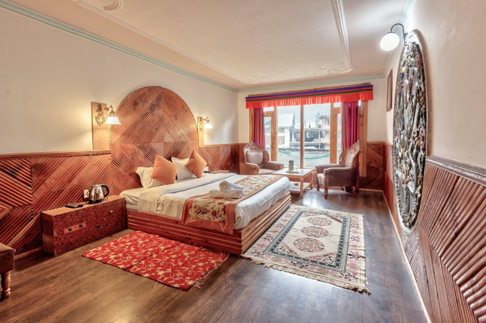 Habitación de hotel con una cama grande, un cabecero de madera, un rincón de descanso con sillones, una mesa de centro y vistas a la ciudad desde la ventana.