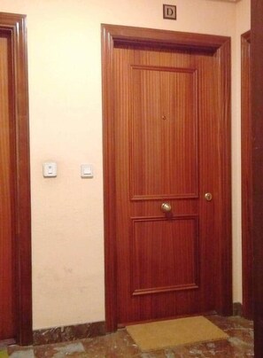 A wooden door with a brass handle and a small brass doorknob, a light switch, and a doormat on a tiled floor.