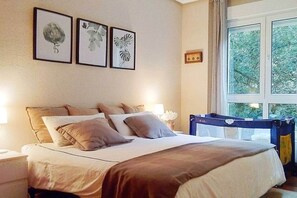 A bedroom with a bed, pillows, a nightstand, a lamp, framed artwork on the wall, and a window with a view of trees.
