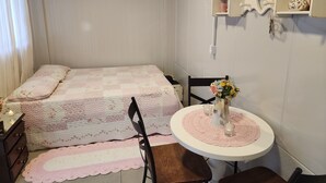 Quarto com cama, uma cadeira, uma mesinha com um vaso e uma estante com objetos.