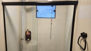 Um chuveiro com paredes de vidro, um chuveiro manual, um espelho de mão e um pequeno armário com produtos de higiene pessoal.