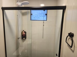 Um chuveiro com paredes de vidro, um chuveiro manual, um espelho de mão e um pequeno armário com produtos de higiene pessoal.