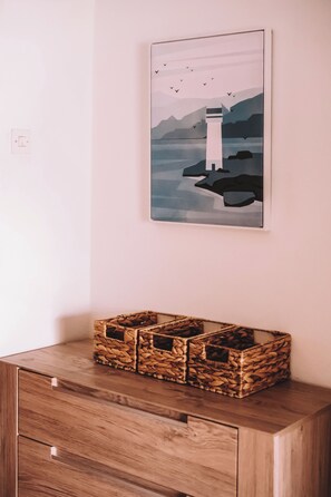 Ein an der Wand befestigtes Kunstwerk, das einen Leuchtturm zeigt, ein Holzkabinett mit Schubladen und ein gespannter Korb darauf.
