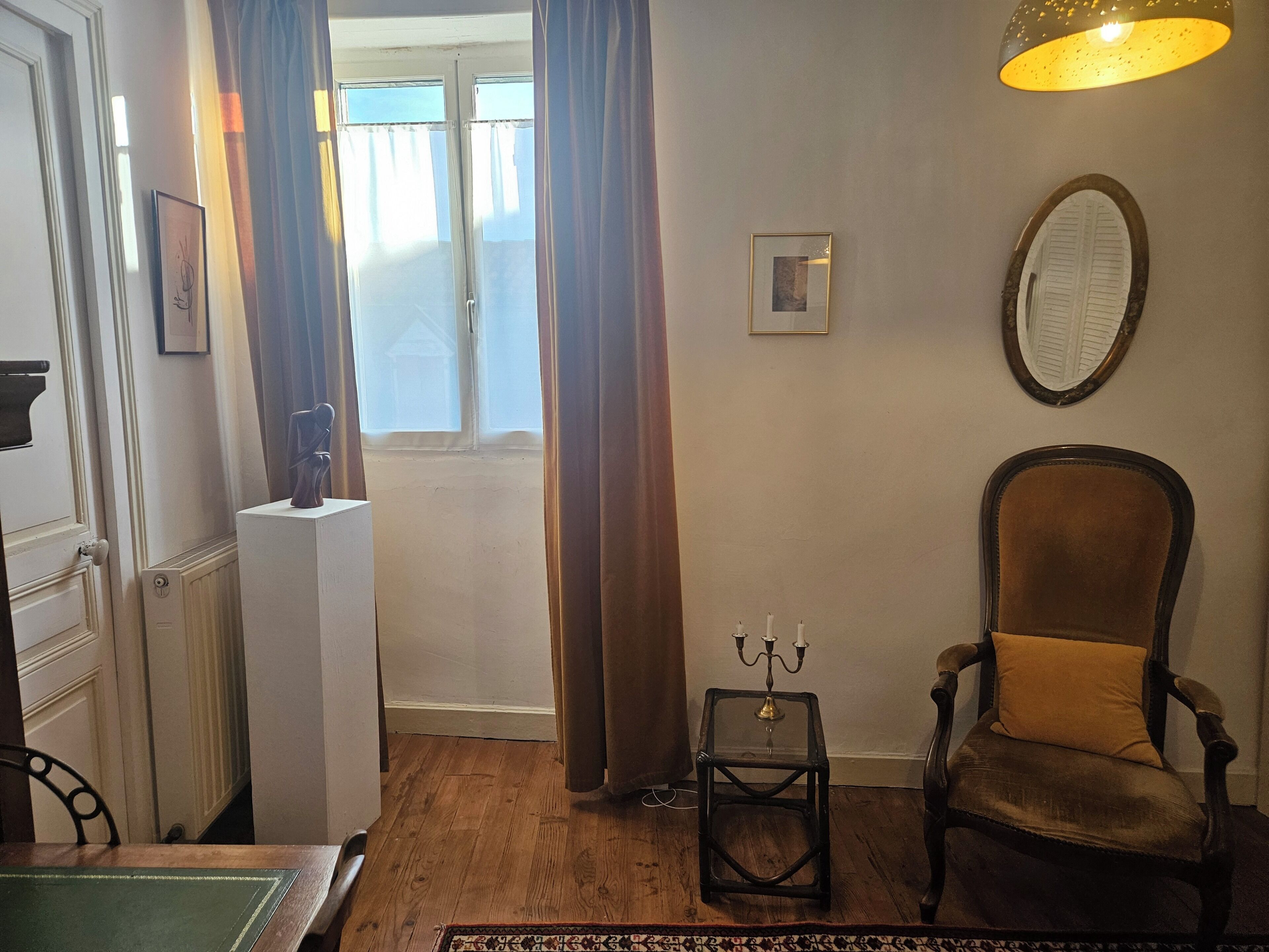 Une pièce avec un plancher en bois, une chaise, une petite table sur laquelle se trouve un chandelier, un radiateur et une fenêtre avec des rideaux.